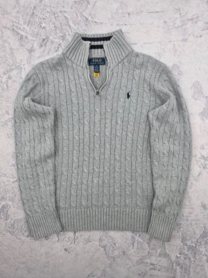 Ljusgrå kabelstickad tröja Polo Ralph Lauren - Ljusgrå kabelstickad tröja från Polo Ralph Lauren med halv dragkedja och hög krage. Tröjan har ribbade muddar och nederkant samt den klassiska broderade loggan på bröstet. Perfekt för lager på lager och riktigt snygg till jeans.