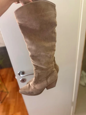 Beiga höga mockastövlar med klack - Snygga höga stövlar i beige mocka med bred klack och rundad tå. Stövlarna har en slouchy passform och är perfekta för att lyfta din outfit. Passar till både jeans och kjol för en trendig look.