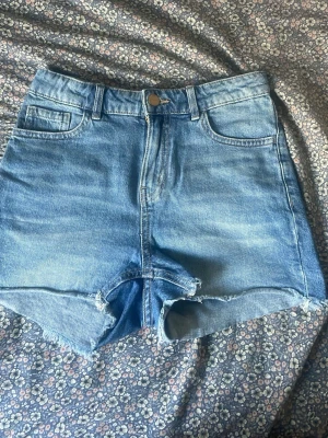 Blå denim shorts med rå kant - Säljer ett par klassiska blå denim shorts med hög midja och rå, fransig kant nedtill. Shortsen har fem fickor, bälteshällor och knappstängning framtill. Perfekta för sommarens alla äventyr!