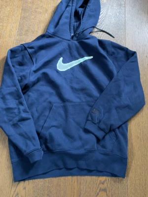Mörkblå Nike hoodie med logga - Mörkblå hoodie från Nike med stor ljusblå Swoosh-logga på bröstet och klassisk känguruficka. Tröjan har huva med dragsko och ribbade muddar vid ärmar och nederkant. Perfekt för en avslappnad och sportig stil. Helt ny använd 3-4 gånger bara 