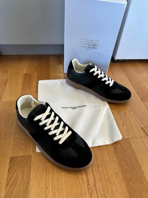 Maison Margiela svarta sneakers - Maison Margiela sneakers i svart mocka och läder med vita skosnören och beige innersula. Klassisk låg modell med gummisula i brunt. Snygg och stilren design som passar till det mesta. Levereras med dustbag och originalkartong.