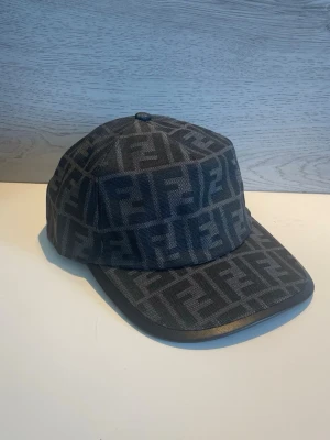 Svart keps med Fendi-logga - Snygg svart keps från Fendi med det klassiska FF-monogrammet över hela kepsen. Tillverkad i tyg med böjd skärm och justerbar passform. Perfekt accessoar för dig som gillar exklusiv streetwear och vill sticka ut med en ikonisk design.