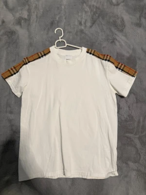 Vit Burberry t-shirt med rutiga axlar - Snygg vit t-shirt från Burberry med klassiska beige-rutiga detaljer på axlarna. T-shirten har rund hals och korta ärmar. Perfekt för dig som vill ha en stilren look med en touch av lyx. Tillverkad i mjuk bomull.                                    Använd 1 gång 
