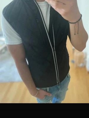 Svart dunväst från Riley, stl S - Mörkblå dunväst från Riley med quiltad design och dragkedja framtill. Märket är etablerat 1992 och västen har en stilren look som passar till jeans och t-shirt. Materialet är dun och västen har en smal passform.