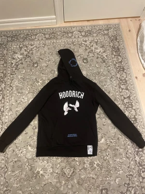 Svart HOODRICH hoodie med duvor - Svart hoodie från HOODRICH med vit text och tryck av två duvor på bröstet och ryggen. På huvan finns blå brodyr och på ryggen står det 'From Nothing To Something' i blått. Klassisk känguruficka och ribbade muddar. Snygg streetwear vibe. I storlek S men kan passa M