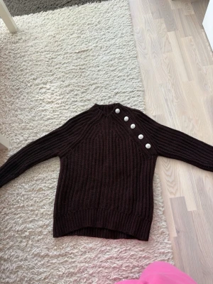 zadig voltaire tröja - Säljer en mörkbrun stickad tröja från Zadig & Voltaire med grov ribbstickning och fem dekorativa knappar i silver längs axeln, perfekt för kyliga dagar.