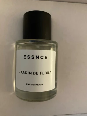 ESSNCE Jardin de Flora parfym - Minimalistisk parfymflaska från ESSNCE med doften Jardin de Flora. Flaskan är cylindrisk i genomskinligt glas med svart rund kork och vit etikett med svart text. Eau de Parfum som ger en fräsch och blommig känsla.