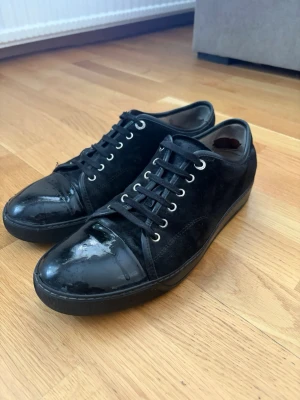 Svarta lanvins - Snygga svarta sneakers från Lanvin med ovandel i mocka och detaljer i blankt läder på tån. Skorna har svarta skosnören, silverfärgade öljetter och en klassisk låg siluett. Perfekta för dig som gillar stilrena och exklusiva sneakers.