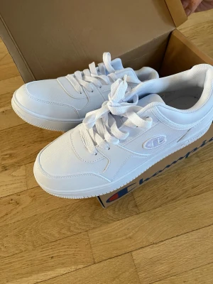 Vita sneakers från Champion - Stilrena helvita sneakers från Champion med klassisk låg profil och tjock sula. Skorna har snörning, diskret logga på sidan och är tillverkade i syntetmaterial och textil. Perfekta för dig som gillar en clean och sportig look.