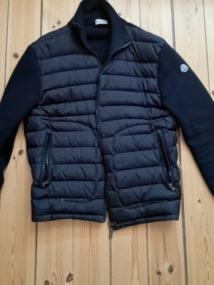 Svart cardiganjacka från Moncler - Snygg svart cardiganjacka från Moncler med quiltad framsida och stickade ärmar. Jackan har två dragkedjefickor framtill och Moncler-logga på ena ärmen. Perfekt för lager-på-lager och streetstyle.