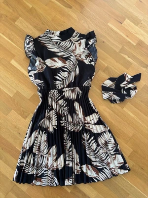 Klänning - Elegant klänning med ett stilfullt lövmönster i svart, vitt och brunt. Modellen har en markerad midja, plisserad kjol och söta volangärmar. Inkluderar matchande skärp.