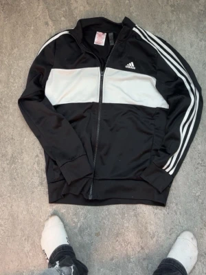 Svart och vit Adidas träningsjacka - Säljer en snygg svart träningsjacka från Adidas med vit panel över bröstet och klassiska vita ränder längs ärmarna. Jackan har dragkedja framtill, ribbade muddar och är tillverkad i ett sportigt syntetmaterial. Perfekt för träning eller chill.