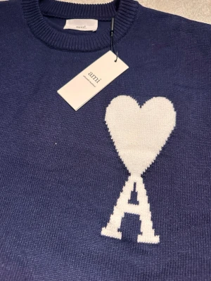 Blå stickad tröja från Ami Paris - Säljer en blå stickad tröja från Ami Paris med ett stort vitt hjärta och bokstaven A på bröstet. Tröjan har rund halsringning, ribbade muddar och är tillverkad i mjukt material. Perfekt för dig som gillar stilrena detaljer och ikoniska loggor. Skriv om du vill snacka om priset!