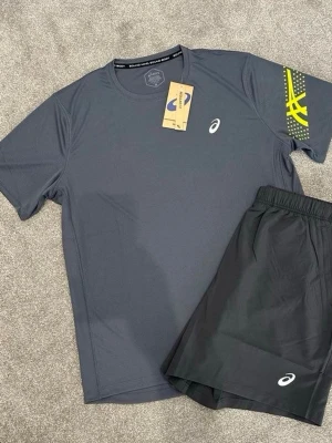 Grå Asics träningsset med tryck - Snyggt träningsset från Asics med en grå t-shirt med gul grafisk detalj på ärmen och matchande svarta shorts. Båda plaggen har Asics-logga och är gjorda i ett lätt, snabbtorkande material som passar perfekt för löpning eller gym.