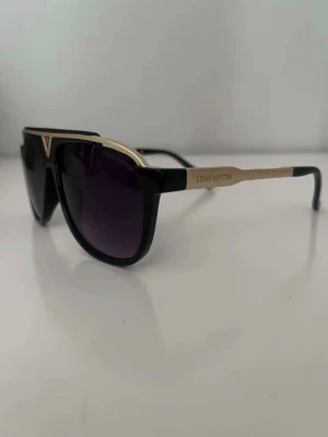 svarta solglasögon - Snygga svarta solglasögon från med guldiga detaljer och mörka glas. Modellen har en modern, kantig form och logotyp på skalmarna. Perfekt accessoar för att lyfta din stil och ge en lyxig touch till outfiten.