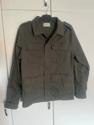 Olivgrön utilityjacka från Stylein - Snygg olivgrön utilityjacka från Stylein med fyra stora fickor framtill, axelklaffar och klassisk krage. Jackan har raka ärmar med knäppning vid ärmslut och är tillverkad i ett slitstarkt material. Perfekt för en cool och avslappnad look.