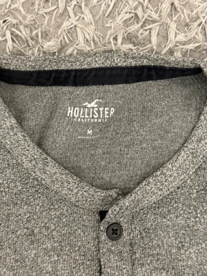 Grå långärmad tröja Hollister - Snygg grå långärmad tröja från Hollister med rund halsringning och knappslå med fyra knappar framtill. Tröjan har en mjuk, melerad struktur och är perfekt för en avslappnad stil. Passar till både jeans och chinos.
