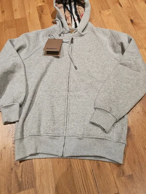 Grå hoodie från Burberry med dragkedja - Snygg grå hoodie från Burberry med hel dragkedja och klassiskt rutigt mönster i huvan. Tillverkad i mjuk bomull och har ribbade muddar samt justerbar huva med snörning. Perfekt för en chill och stilren look. Storlek L men liten i storleken, mer som en S. Helt ny, aldrig använd.