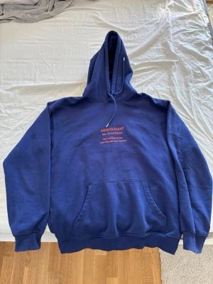 Blå hoodie från Valient med tryck - Mörkblå hoodie från Valient med röd texttryck på bröstet där det står 'AMBIVALENT' och en definition. Klassisk känguruficka framtill, ribbade muddar och justerbar huva med dragsko. Perfekt för en chill och avslappnad stil.