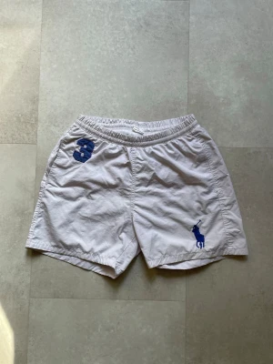 Vita badshorts från Polo Ralph Lauren - Vita badshorts från Polo Ralph Lauren med blå logga och siffran 3 broderad på vänster sida. Shortsen har elastisk midja och är tillverkade i ett lätt bomullsmaterial. Perfekta för varma dagar och enkel att matcha med olika toppar.