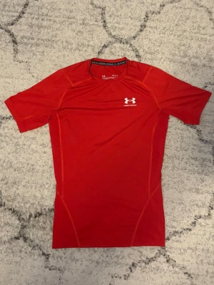 Röd träningströja från Under Armour - Snygg röd träningströja från Under Armour med korta ärmar och tight passform. Tillverkad i ett lätt och stretchigt funktionsmaterial som transporterar bort fukt. Logga i vitt på bröstet och nacken. Perfekt för gymmet eller löpning.