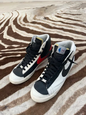 Nike Blazer Mid 77 NBA 75th Anniversary - Nike Blazer Mid 77 sneakers med svart och vitt läder, svarta skosnören och kontrasterande Swoosh-loggor i rött och svart. NBA 75th Anniversary-detaljer på plösen och hälen. Klassisk retrostil med högt skaft och robust sula.