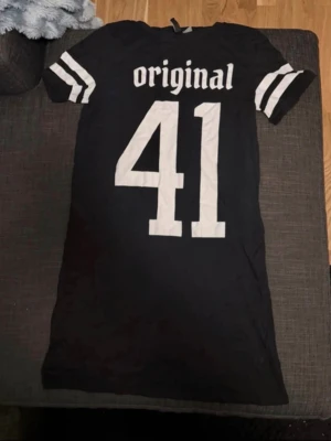 Svart t-shirtklänning med tryck - Cool svart t-shirtklänning med vita tryck, texten 'original 41' fram och 'so over it' bak. Sportiga ränder på ärmarna ger en avslappnad vibe. Perfekt för dig som gillar streetstyle och vill ha något bekvämt och trendigt.