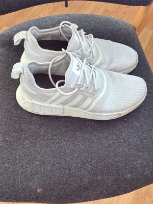 Adidas NMD vita sneakers - Säljer ett par vita Adidas NMD sneakers med klassiska tre ränder på sidan. Skorna har mesh-överdel, snörning och en bekväm Boost-sula. Perfekt för dig som gillar sportig stil och vill ha något lätt och snyggt på fötterna.
