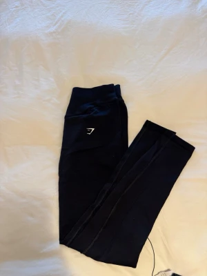 Svarta leggings från Gymshark - Säljer ett par svarta leggings från Gymshark med hög midja och diskret logga på höften. Tillverkade i stretchigt och mjukt material som sitter skönt på kroppen. Perfekta för träning eller chill. Tight passform och snygg design.