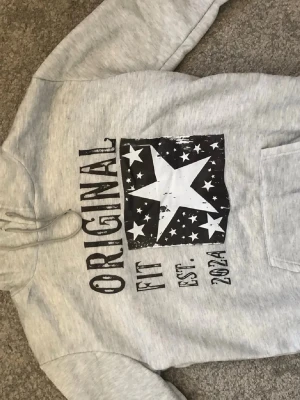 Grå hoodie med stjärntryck - Snygg ljusgrå hoodie med stor vit stjärna och svart tryck framtill där det står 'ORIGINAL FIT EST. 2021'. Hoodien har huva med dragsko och en klassisk magficka. Perfekt för en avslappnad streetwear-look. Passar även M