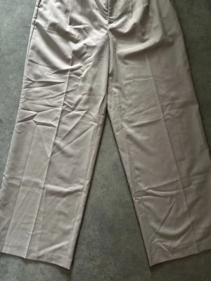 Beige vida kostymbyxor från Gina Tricot - Snygga beige kostymbyxor från Gina Tricot i storlek 34. Byxorna har en rak och vid passform med pressveck framtill och hällor i midjan. Perfekta att styla med en skjorta eller crop top. Materialet är mjukt och har en lätt glans.