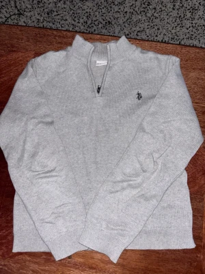 Ljusgrå halvzip tröja från U.S. Polo Assn. - Ljusgrå stickad tröja från U.S. Polo Assn. med halv dragkedja och broderad logga på bröstet. Tröjan har ribbade muddar vid ärmslut och nederkant samt låg krage. Perfekt för lager-på-lager och enkel att matcha. Den passar storlek 15-16 och även M.