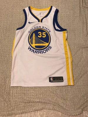 Golden State Warriors basketlinne - Golden State Warriors basketlinne med nummer 35 och Durant på ryggen. Linnet är vitt med blå och gula detaljer, har rund hals och ärmlös design. Tillverkat i lätt och ventilerande polyester, perfekt för basket eller träning.