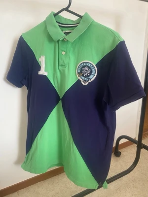 Hackett Grön och marinblå pikétröja med nummer - Säljer en cool pikétröja från Hackett London i grön och marinblå färg med stora diagonala fält. Tröjan har klassisk krage, knappar framtill och ett broderat märke på bröstet samt siffran 1 både fram och bak. Perfekt för dig som gillar sportig stil.