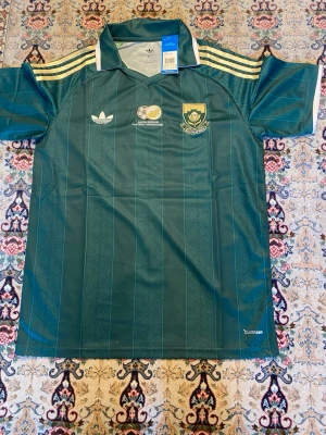Grön Adidas fotbollströja Sydafrika - Säljer en grön fotbollströja från Adidas med Sydafrikas fotbollsförbunds emblem på bröstet. Tröjan har klassiska Adidas-ränder i gult på axlarna, vit krage och diskret mönster i tyget. Perfekt för dig som gillar retro och sport.