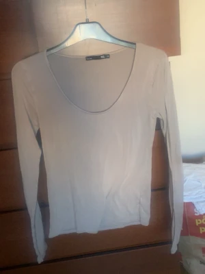 Beige långärmad topp Gina Tricot - En enkel och stilren beige långärmad topp från kafé 157.. Toppen har en djup rundringning och är tillverkad i ett mjukt, stretchigt material som sitter skönt mot kroppen. Perfekt att matcha med jeans eller kjol för en clean look.