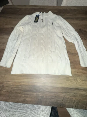 Vit kabelstickad tröja från Polo Ralph Lauren - Kabelstickad vit tröja från Polo Ralph Lauren med halv dragkedja och broderad logga på bröstet. Tröjan har ribbade muddar och nederkant, samt en klassisk hög krage. Perfekt för dig som gillar stilrena och tidlösa plagg.