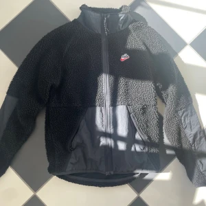 Svart teddyjacka från Nike - Mysig svart teddyjacka från Nike med dragkedja framtill och höga fickor. Jackan har kontrastmaterial på ärmar och nedre delen samt en liten Nike-logga på bröstet. Perfekt för kalla dagar och riktigt skön att bära. Trendig! 