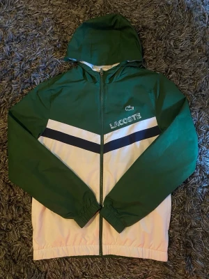 Grön och vit vindjacka från Lacoste - Snygg vindjacka från Lacoste i grönt och vitt med svarta detaljer. Jackan har huva, dragkedja framtill och elastiska muddar. Stora Lacoste-loggan och krokodilen på bröstet. Perfekt för en sportig och trendig look.