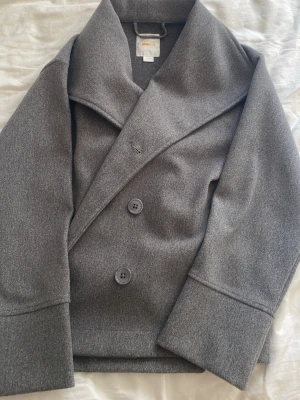 Grå skepparkavaj från Gina Tricot - Stilren grå skepparkavaj från Gina Tricot med bred krage och dubbelknäppning framtill. Kavajen har raka ärmar och är tillverkad i ett mjukt tyg som ger en klassisk look. Perfekt för dig som vill ha en tidlös jacka med snygg passform.