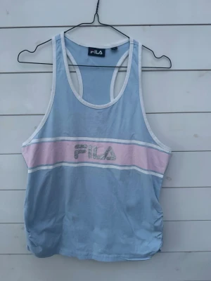 Blå och rosa linnetopp från FILA - Snygg blå linnetopp från FILA med breda axelband och vit kant. Toppen har en pastellrosa rand över bröstet med FILA-loggan i silver. Tillverkad i mjuk bomull, perfekt för en sportig och avslappnad look.