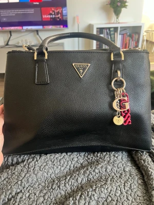 Svart handväska från Guess - Snygg svart handväska från Guess i skinnimitation med guldfärgade detaljer och dekorativa Guess-berlocker. Väskan har två handtag, dragkedja upptill och flera innerfack för smart förvaring. Perfekt accessoar för att lyfta din outfit.