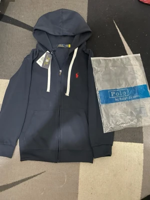Mörkblå hoodie från Polo Ralph Lauren - Snygg mörkblå hoodie från Polo Ralph Lauren med dragkedja, vit dragsko och klassisk röd logga på bröstet. Hoodien har känguruficka och ribbade muddar. Perfekt för en chill och stilren look.