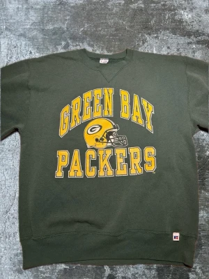 Green Bay Packers sweatshirt - Mörkgrön sweatshirt med stort gult Green Bay Packers-tryck och hjälm på bröstet. Klassisk crewneck-modell med ribbade muddar och avslappnad passform. Perfekt för dig som gillar amerikansk fotboll och sportig stil.
