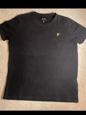 Svart t-shirt från Lyle & Scott - Svart t-shirt från Lyle & Scott med klassisk rund hals och korta ärmar. T-shirten har en gul broderad logga på bröstet och är tillverkad i mjuk bomull. Perfekt för en clean och stilren look.