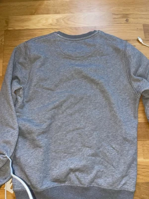 Grå sweatshirt från GANT - En klassisk grå sweatshirt från GANT i mjukt bomullsmaterial. Tröjan har rund halsringning, ribbade muddar vid ärmslut och nederkant. Perfekt för en avslappnad och stilren look.