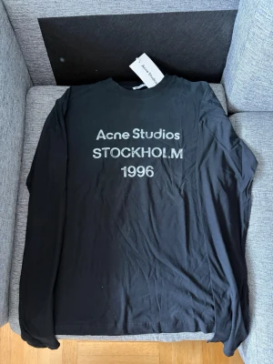Acne studios long sleeve - Svart långärmad t-shirt från Acne Studios med trycket 'Acne Studios STOCKHOLM 1996' i vitt framtill. Klassisk rund hals och relaxed fit. Perfekt för dig som gillar stilrena och trendiga plagg med streetkänsla.