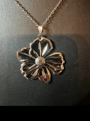 Silverfärgat halsband med blomma - Ett snyggt halsband med en stor, silverfärgad blomma som hänge. Blomman har en detaljerad och öppen design med markerad mitt och vågiga kronblad. Kedjan är tunn och elegant, perfekt för att ge ett statement till din outfit.