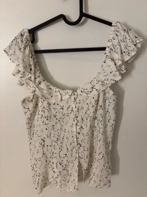 Vit blommig blus med volang - Söt vit blus med svart blommönster och bred volang runt halsen. Blusen är ärmlös och har en luftig, lätt form som passar perfekt till jeans eller kjol. Materialet är tunt och flowy, vilket ger en skön känsla. Ingen synlig logga eller märke. Säljs då jag inte använder den.