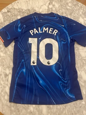 Chelsea matchtröja Palmer #10 Nike - Säljer en blå Chelsea matchtröja från Nike med Palmer och nummer 10 på ryggen. Tröjan har ett unikt mönster i olika blå nyanser, klubbmärke på bröstet och Premier League-logga på siffrorna. Perfekt för fotbollsfans!